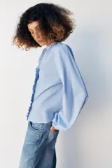 Blusa celeste de popelín, con cuello subido y escote de pico, manga larga abullonada terminada en elástico, y detalles de volantes en el cuello y la botonadura frontal con cierre de botones ocultos.
