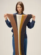Bufanda tejida en punto acanalado con diseño de franjas verticales en tonos tierra, incluyendo marrón, beige, mostaza y negro con motas. Presenta una etiqueta metálica pequeña en uno de sus extremos.