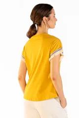 Remera de manga corta color mostaza, confeccionada en 100% algodón, con cuello redondo y detalles de flecos y guardas geométricas en las mangas.