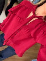 Camisa de color rojo intenso con volados en los hombros y lazo ajustable en el cuello. Presenta un cierre frontal con botones y un diseño de calce amplio.