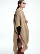 Ruana tejida de color beige con capucha integrada y terminaciones con flecos en el ruedo. Presenta un diseño envolvente y holgado, confeccionada en una mezcla de lana merino extrafina y viscosa.