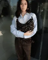 Bolso de hombro tipo hobo, confeccionado en gamuza color marrón oscuro, decorado con tachas metálicas plateadas en toda su superficie frontal. Cuenta con un asa corta fija y cierre superior.