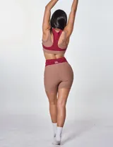 Conjunto deportivo de dos piezas compuesto por un top y un short de tiro alto. El top es de estilo deportivo con doble bretel y diseño bicolor en tonos bordó y marrón, con el logo 'bk.' estampado en el frente. El short es de corte ajustado, con diseño acanalado y paneles laterales en contraste de color bordó.