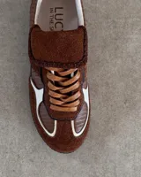 Championes de cuero vacuno con diseño de paneles en tonos marrones y detalles en blanco. Presentan una combinación de texturas lisas y grabadas, con cordones planos y costuras decorativas en la lengüeta.