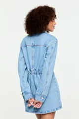 Vestido camisero corto confeccionado en denim azul claro, con cuello camisero, cierre frontal de botones metálicos, bolsillos frontales con solapa en el pecho y diseño entallado en la cintura.