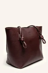 Cartera tote de cuero suave color marrón, con diseño amplio y líneas simples, con tiras de hombro decoradas con nudos artesanales y pequeño logo grabado en el centro.