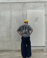 Remera oversize gris oscuro con corte holgado, mangas cortas y estampa trasera en blanco y negro con forma de estampilla que muestra a una persona con un gato y el texto 'Kaboa Industries'.