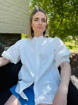 Camisa blanca de popelina de algodón, manga corta abullonada y cuello mao. Presenta doble detalle de puntilla en forma de V en la parte superior delantera.