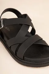 Sandalia baja de tiras cruzadas color camel, con hebilla metálica ajustable en el tobillo y suela negra gruesa.