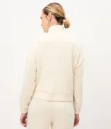 Buzo deportivo de microfibra color beige claro, con cuello alto, cierre frontal parcial y manga larga. Presenta recortes laterales en las mangas con bandas en celeste y líneas negras.