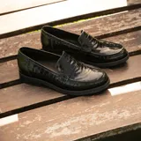 Mocasín de cuero vacuno negro con textura grabada tipo reptil. Presenta un diseño clásico con antifaz en el empeine, suela de goma en tono natural y taco bajo de madera.