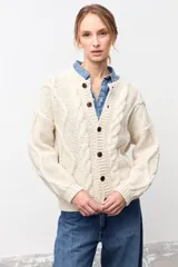 Cardigan de punto grueso color crudo, con diseño de trenzas verticales, cuello redondo y cierre frontal con botones oscuros.