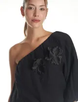 Blusa de un hombro color negro, con diseño asimétrico, una manga larga abullonada, forro interno y apliques de flores de tela en el frente.