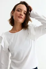 Remera de manga larga en color crudo, confeccionada en tejido acanalado, con detalle de volados en los hombros y cuello redondo.