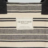 Bolso tipo shopper de tela color crudo con rayas horizontales negras. Tiene doble asa corta negra y etiqueta rectangular con texto en el centro.