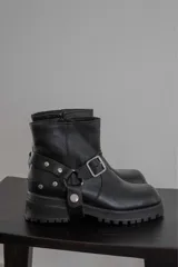 Bota corta de cuero negro con punta cuadrada, suela track y detalle de estribo desmontable con hebilla metálica.