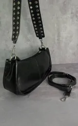 Cartera tipo baguette de cuero sintético negro con acabado brillante, cierre superior y correa ancha decorada con tachas metálicas plateadas.