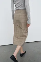 Falda midi de tiro medio con estampado de cuadros en tonos beige y gris. Presenta un diseño cruzado con un volante decorativo en el delantero y cierre con botón.