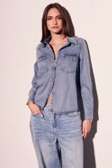 Camisa de denim celeste con cuello clásico, cierre frontal con botones a presión, dos bolsillos con solapa abotonada en el pecho y mangas largas con puños abotonados.