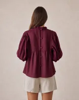 Blusa holgada color bordó con detalle de nido de abeja en el canesú delantero, cuello alto con volados y mangas tres cuartos con puños elásticos fruncidos.