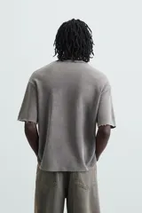 Camiseta gris de punto con efecto lavado y roturas, corte "boxy fit", cuello redondo y manga corta. Presenta un estampado frontal en blanco y rojo con diseño abstracto.