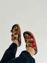 Sandalias de cuero marrón con tres tiras ajustables con hebillas doradas y detalles en rojo.