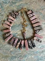 Collar de rodocrosita natural compuesto por piezas rectangulares en tonos rosa, lila y negro, unidas con detalles metálicos dorados y cadena de extensión con dije colgante.