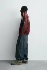 Cazadora de tejido técnico color bordó, corte relajado, con cuello subido y cierre frontal de cremallera. Presenta paneles en color negro en los laterales y detalles de ribete en color gris claro y blanco que recorren los hombros y mangas.