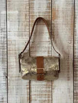 Cartera tipo mini bag de cuero con acabado metalizado dorado, solapa frontal con detalle de correa y hebilla central en cuero marrón, y correa de hombro ajustable.