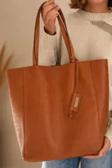 Tote bag confeccionada 100% en cuero vacuno color suela, con cierre de broche imán, sin forro interior y bolsillo interno con cierre. Incluye tiras largas para el hombro y detalle colgante con placa metálica grabada.
