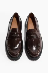 Mocasines tipo penny de color marrón oscuro, confeccionados en material sintético con acabado brillante. Presentan flecos decorativos en el empeine, una banda transversal con detalle metálico y una suela gruesa dentada de color negro.