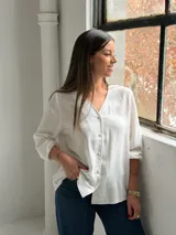 Blusa blanca de manga larga con cuello en V y cierre frontal de botones. Presenta una textura sutil en la tela y un corte recto y holgado.