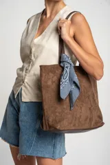 Bolso tipo tote de gran tamaño en color camel, confeccionado en cuero vegano con acabado efecto gamuza. Presenta doble asa larga para llevar al hombro y una placa metálica pequeña con el logo de la marca en el frente.
