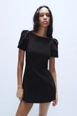 Vestido corto de color negro con cuello redondo y mangas cortas abullonadas. Presenta un diseño entallado con costuras frontales y cierre de cremallera oculto en la espalda.