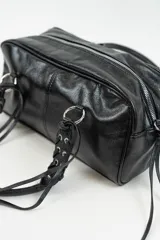 Bolso de mano tipo bowling de cuero sintético negro, con diseño estructurado, costuras decorativas y detalles de tiras largas en los laterales. Cuenta con asas cortas trenzadas con argollas metálicas.