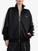 Chaqueta de estilo deportivo y corte oversize, color negro con franjas blancas en las mangas y cuello alto. Cuenta con cierre frontal completo de cremallera y bolsillos laterales.