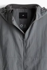 Campera rompevientos de nylon color gris oscuro, con forro de malla. Presenta cierre frontal con protección para el mentón, capucha ajustable con cordón, bolsillos laterales y un bolsillo interior, todos con cierre. Los puños y el bajo tienen elástico revestido.