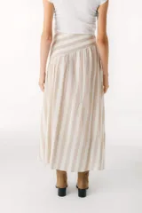 Falda larga color beige de lino, con corte amplio y vuelo, que presenta un canesú diagonal en la parte superior que genera volumen fruncido en la falda.