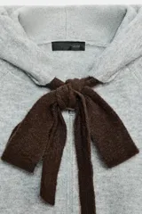 Chaqueta de punto con capucha, manga larga y cierre frontal con cremallera. Presenta un diseño en color beige con un lazo decorativo a contraste en color marrón oscuro en la zona del cuello.