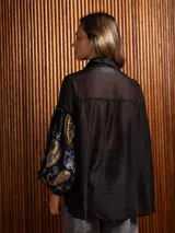 Camisa de manga larga con diseño de terciopelo estampado con motivos florales y paisley, combinada con paneles de tela transparente en la parte superior. Presenta cuello clásico, cierre frontal con botones y mangas abullonadas.
