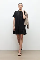 Vestido corto negro de corte amplio, con cuello mao, mangas cortas con volados y detalles de vainillas verticales en el frente. Cuenta con un volante fruncido en el ruedo.