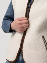 Chaleco de polar tipo corderito en color beige, con cierre frontal y detalles de ribetes en color marrón. Presenta bolsillos laterales y un diseño sin mangas.