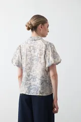 Camisa de manga corta abullonada, con cuello camisero y botones frontales. Presenta un estampado de inspiración barroca en tonos marrón y beige claro.