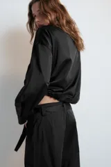 Cazadora satinada de color negro, con cuello subido, manga larga y bolsillos frontales de plastrón con cordones decorativos. Cuenta con bajo elástico y cierre frontal con cremallera y botones a presión oculta.