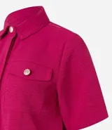 Camisa color fucsia de tejido airflow, con cuello camisero, mangas cortas con pliegues, cierre frontal con botones metalizados y bolsillos con solapa en el pecho.