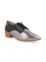 Zapato acordonado de estilo oxford con diseño bicolor. Presenta una puntera y lateral en cuero metalizado plateado, mientras que la parte trasera y el área de los cordones están confeccionados en cuero con textura de reptil en color negro. Incluye un taco bajo de madera y suela a tono.