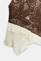 Top sin mangas con cuello redondo, confeccionado en una combinación de tejido de crochet marrón con diseño floral y base de tela color crudo con volados asimétricos y detalles de puntilla. Cuenta con cierre de botón en la espalda.