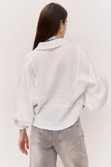 Camisa blanca de corte amplio y largo cropped, con mangas abullonadas, puño ajustado, cuello clásico y abertura en V con botones al tono.