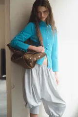 Bolso tipo shopper de hombro, confeccionado en gamuza color marrón, decorado con tachas metálicas plateadas en toda su superficie. Cuenta con doble asa de hombro y cierre mediante botón imantado.