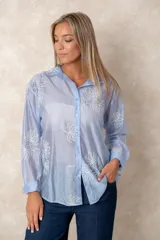 Camisa de manga larga en tono celeste traslúcido, con cuello clásico y cierre frontal de botones. Presenta un delicado bordado de motivos florales en hilo blanco distribuido por toda la prenda.